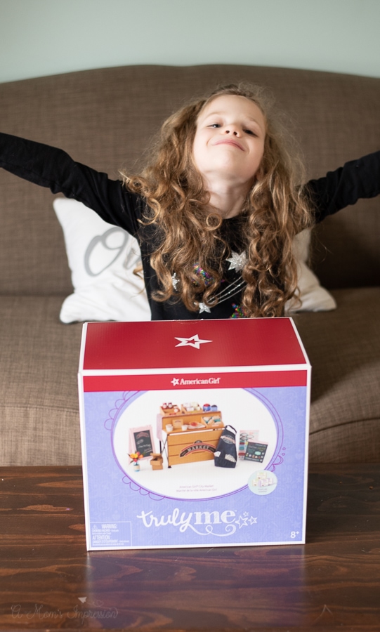 Girl spreading her arms in excitement with a box that includes am american girl playset in it. 