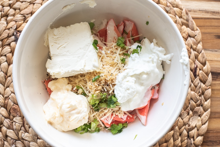 A white bowl with imitation crab, cream cheese, mayo, sour cream, green onions and parmesan cheese in it. 