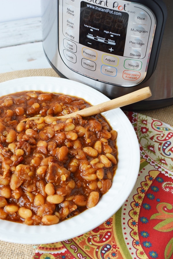 a bowl of baked beans sitting in front of a pressure cooker on a table with a wooden spoon