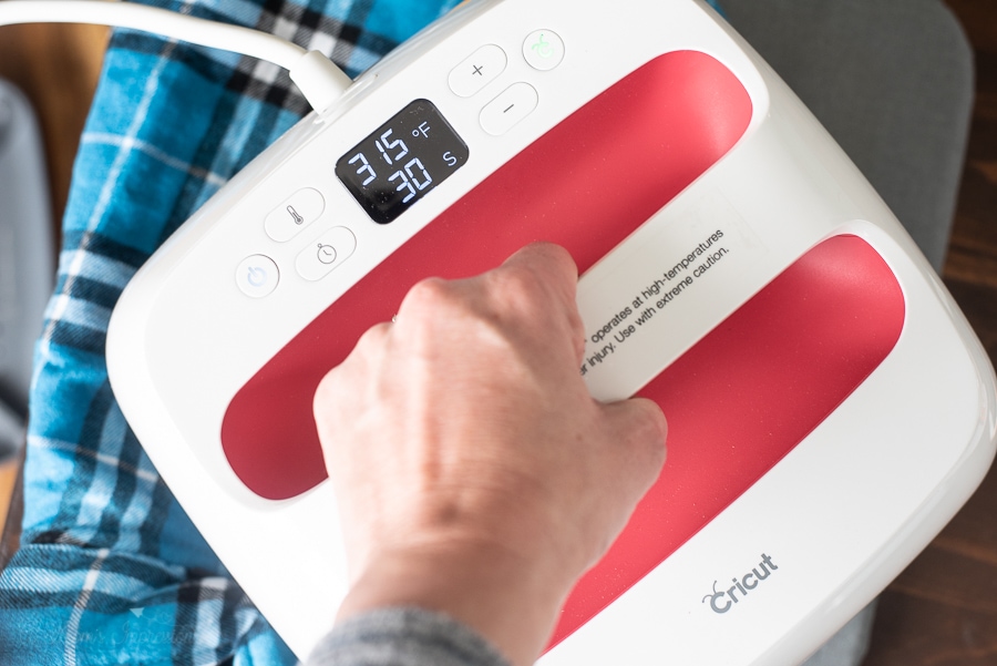 A person using a Cricut EasyPress 2 to iron on a decal to a blue plaid dog jacket